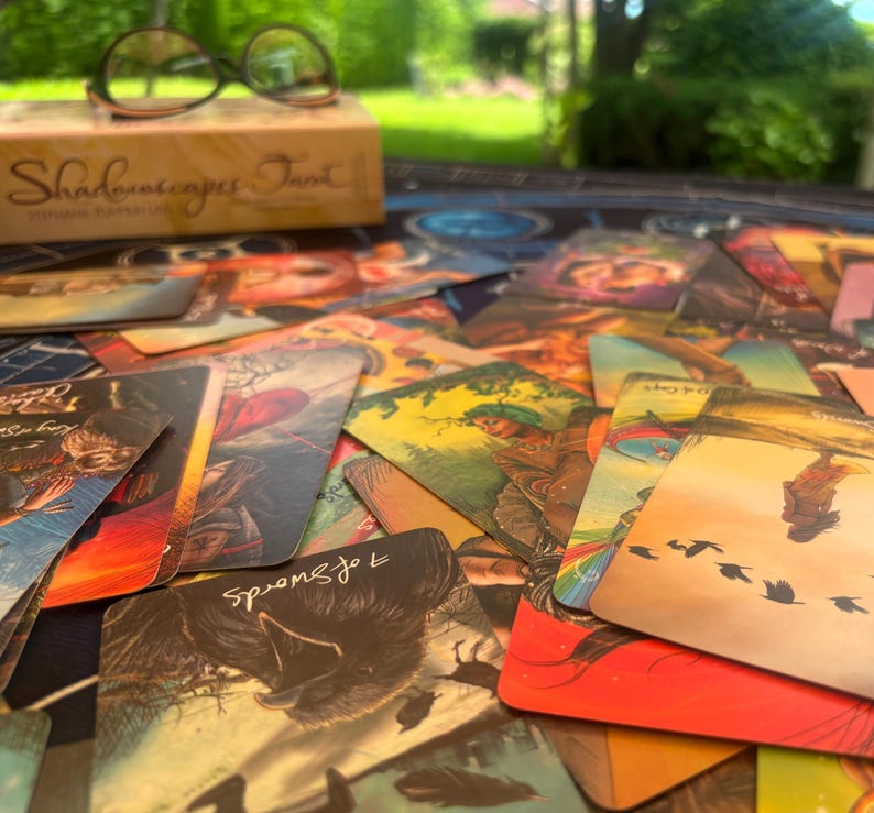 May include: A collection of Shadowscapes Tarot cards spread out on a surface, featuring colorful illustrations of figures, animals, and nature scenes. A box labeled Shadowscapes Tarot and a pair of black-rimmed glasses sit in the background.