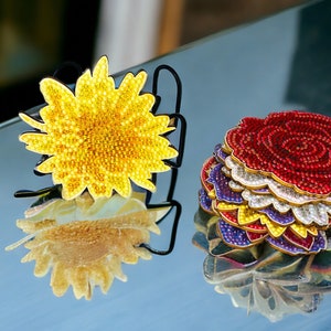 May include: Two decorative flower brooches, one yellow and one red, with a layered design. The yellow brooch is a sunflower with a beaded center and the red brooch is a rose with a beaded center.