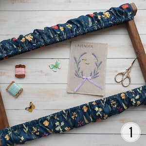 Può includere: Un tessuto floreale blu e bianco teso su un telaio da ricamo in legno. Il telaio contiene un pezzo di tessuto di lino con una ghirlanda di lavanda ricamata e la parola "Lavender" in alto. Ci sono tre api ricamate nella ghirlanda. Ci sono anche due farfalle, un rocchetto di filo e un paio di forbici sulla superficie di legno bianco.