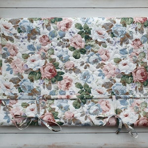 Peut inclure: Tissu floral matelassé avec un motif de roses roses, bleues et blanches. Le tissu est fixé à une tringle en bois et comporte des liens en bas. Le fond est une surface en bois blanc.