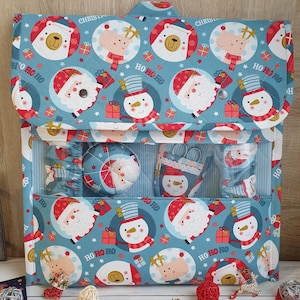 May include: A blue Christmas-themed organizer with a flap closure, featuring a pattern of Santa Claus, snowmen, reindeer, and bears. The organizer contains sewing supplies and accessories. The text "HO HO HO" is visible.