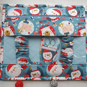 May include: A Christmas-themed sewing organizer with a blue background and a pattern of Santa Claus, snowmen, and gift boxes. The organizer has a zippered pocket and several compartments, including one with scissors. The words "HO HO HO" are printed on the fabric.