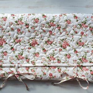 May include: A white quilted fabric with a floral pattern in shades of pink. The fabric is rolled up and tied with two white ribbons. The fabric is on a white wooden surface.