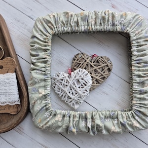 Pode incluir: Uma moldura quadrada de tecido com estampa floral em tons de verde, azul e creme, com borda franzida. Duas decorações de coração tecidas, uma branca e outra marrom, estão dentro da moldura. Uma bandeja de madeira com tesoura e renda ao fundo.