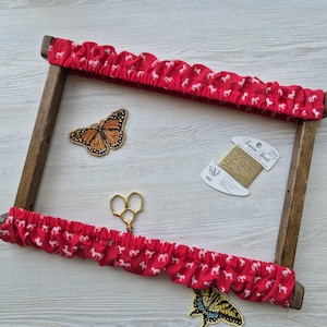 Puede incluir: Un bastidor de bordado de madera con tela roja con siluetas de caballos blancos. El bastidor contiene una mariposa bordada, hilo dorado y unas tijeras doradas pequeñas. También hay otra mariposa.