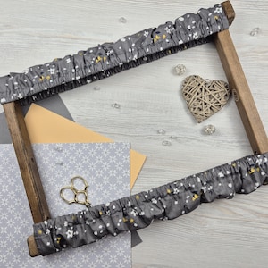 Puede incluir: Un bastidor de bordado de madera con una cubierta de tela floral gris. La tela tiene un patrón de pequeñas flores blancas y amarillas. También se ve una decoración en forma de corazón tejido y unas tijeras.