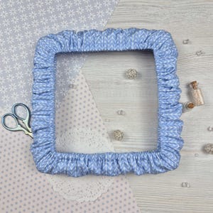 Pode incluir: Uma capa de bastidor de bordado azul claro quadrada com um padrão de chevron branco. O tecido é franzido nas bordas. O fundo inclui tesouras, papel decorativo e pequenos enfeites.