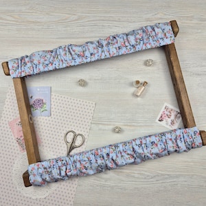 Puede incluir: Un bastidor de bordado de madera con ribete de tela floral azul claro. La tela tiene un estampado de pequeñas flores rosas y rojas. Los suministros de artesanía, incluyendo tijeras y adornos decorativos, rodean el marco.