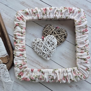 Pode incluir: Uma moldura quadrada com um padrão floral de rosas cor-de-rosa sobre um fundo branco. Duas decorações de coração de vime, uma branca e outra castanha, estão dentro da moldura. A moldura está sobre uma superfície de madeira clara.