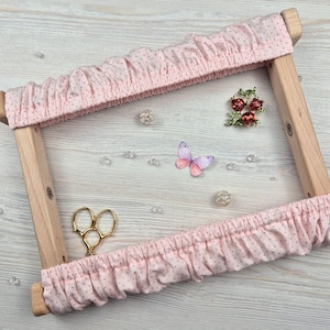 Puede incluir: Un bastidor de bordado de madera con tela rosa salpicada de pequeños círculos rosa oscuro. La tela está fruncida alrededor del marco. Unas tijeras doradas, una mariposa y elementos decorativos están en la superficie.