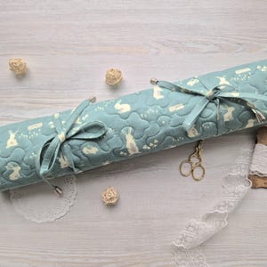 May include: A teal blue fabric roll with a white bunny pattern. The roll is tied with a teal blue ribbon and is sitting on a light wood surface.