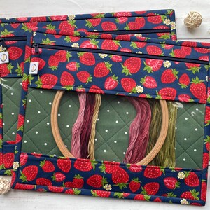 May include: Two rectangular craft pouches with a navy blue strawberry pattern. Each pouch has a clear window, a green quilted interior, and a zipper closure. Embroidery floss and a wooden hoop are visible inside.