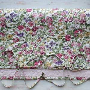 Op de afbeelding: Gewatteerde bloemenstof met een witte achtergrond en een patroon van roze, paarse en gele bloemen. De stof heeft een geschulpte rand met drie strikken. De onderste rand is een lichtroze stof met een stippenpatroon.