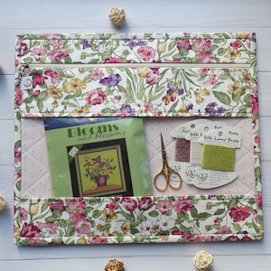 Può includere: Un organizer per artigianato con stampa floreale con cerniera superiore, contenente un kit per punto croce, forbici e filo da ricamo. L'organizer presenta un interno rosa trapuntato e un colorato motivo floreale.