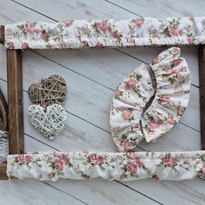 Puede incluir: Un bastidor de bordado de madera con fundas de tela floral en rosa y verde. También se ven dos corazones decorativos tejidos y un ribete de encaje. La tela tiene un fondo blanco con rosas rosas y hojas verdes.