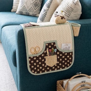 May include: A quilted armrest organizer in cream and brown with a floral pattern, featuring pockets for scissors, thread, and needles. The organizer is designed to fit over the arm of a blue sofa, with a small attached cup holder.