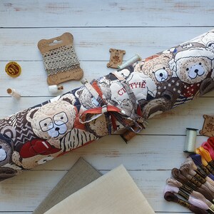 May include: A rolled-up fabric storage case with a brown and white pattern of teddy bears. The case is tied with a red ribbon and has the word "CTYTIE" printed on it. The case is surrounded by various colors of embroidery floss and other sewing supplies.