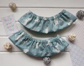 May include: Two blue fabric baby bibs with a white rabbit pattern. The bibs are gathered at the top and have a curved shape. A card with text and decorative balls are also in the image.