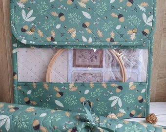 Pode incluir: Um organizador de artesanato verde acolchoado com um padrão de outono de bolotas e folhas. O organizador tem uma janela transparente mostrando bastidores de bordado e tecido. Um tapete enrolado a condizer é amarrado com uma fita.