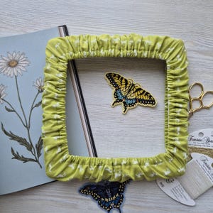Pode incluir: Uma moldura quadrada de tecido verde limão com estampa de animais branca. Um patch de borboleta amarela e preta com detalhes azuis está dentro da moldura. Um caderno com uma ilustração de margarida e tesouras douradas também são visíveis.