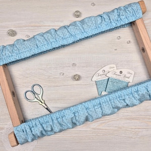 Pode incluir: Um bastidor de bordado de madeira com tecido azul claro com bolinhas pretas. O tecido é franzido ao redor da moldura. Pequenas tesouras e dois carretéis de linha azul também são visíveis.