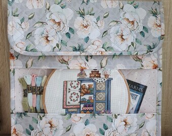 Pode incluir: Uma bolsa de projeto acolchoada cinza com uma estampa floral de rosas brancas. A bolsa tem uma alça e uma janela transparente de plástico que mostra um projeto de ponto cruz, um aro de bordado e fio de bordar.