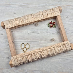 Op de afbeelding: Een houten borduurring met beige stof met kleine zwarte stippen. De ring is rechthoekig en heeft een lichte houten afwerking. Gouden schaar en decoratieve broches zijn ook aanwezig.