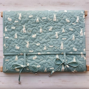 May include: A teal blue quilted baby crib bumper with a white bunny rabbit pattern and white text that says "Little Bunny". The bumper is tied with teal blue ribbons.