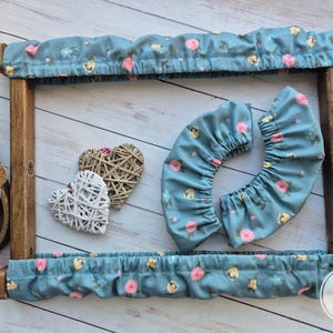 Pode incluir: Um aro de bordado de madeira com uma capa de tecido floral azul. O aro é decorado com dois ornamentos em forma de coração branco e marrom. A capa de tecido tem uma borda franjada e um padrão floral com flores rosa e amarelas. O número "3" é visível no canto inferior direito da imagem.