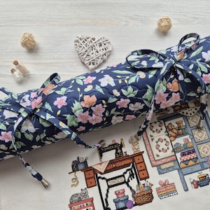 May include: A blue quilted fabric roll with a floral pattern and a white drawstring closure. The roll is lying on a white surface with a cross-stitch pattern of a sewing room.
