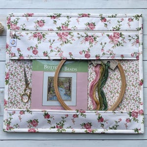 Könnte beinhalten: Ein weiß-rosa Blumenmuster-Bastel-Organizer mit Reißverschluss. Im Inneren befinden sich Stickutensilien, darunter ein Holzrahmen, eine Schere, Stickgarn und ein Kreuzstich-Set mit dem Text "Button Beads".