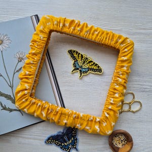 Pode incluir: Uma moldura coberta de tecido amarelo com um padrão de unicórnio branco. Um patch de borboleta amarelo, preto e azul está dentro da moldura. Um caderno azul com uma ilustração de margarida e tesouras douradas também são visíveis.
