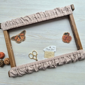 Pode incluir: Um bastidor de bordado de madeira com uma capa de tecido com bolinhas, junto com um enfeite de borboleta e abóboras com contas. Tesouras e linha douradas também estão presentes, sugerindo um tema artesanal.