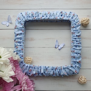 Pode incluir: Uma moldura quadrada de tecido azul claro com um padrão floral de flores vermelhas e azuis. A moldura tem uma borda com babados. Borboletas decorativas e pequenas bolas tecidas são colocadas ao redor da moldura em uma superfície de madeira clara.