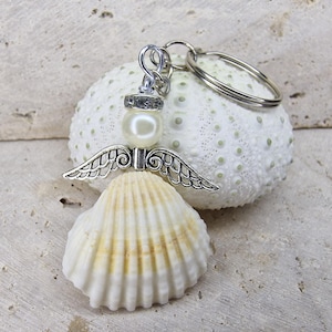 Könnte beinhalten: Ein Muschelangel-Schlüsselanhänger mit Perlenkopf und silberfarbenen Flügeln. Der Körper des Engels ist eine weiße Jakobsmuschel mit gelben Akzenten. Der Schlüsselanhänger ist an einem silberfarbenen Ring befestigt und liegt auf einer weißen, strukturierten Oberfläche.