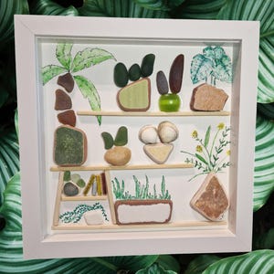 Pode incluir: Uma obra de arte emoldurada em branco com uma prateleira feita à mão de materiais naturais. A prateleira é decorada com vários pedaços de vidro do mar e conchas, criando uma cena de jardim em miniatura. A obra de arte inclui uma palmeira, uma planta monstera e outras plantas verdes, todas feitas de materiais naturais.