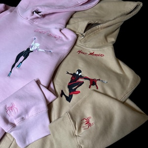 May include: Two hooded sweatshirts with embroidered Spider-Man characters. One is pink with Gwen Stacy and the other is tan with Miles Morales.