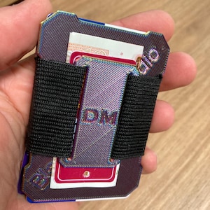 Può includere: Un portafoglio in metallo nero e iridescente con un elastico nero. Il portafoglio ha le lettere "DM" stampate su di esso e la parola "UID" sul lato.