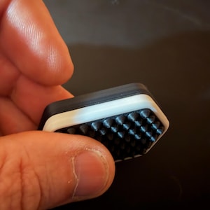 May include: A hexagonal object featuring a black top surface with a grid of small, pointed spikes. The sides of the object are layered with a white middle section sandwiched between a black top and a black base, creating a distinct three-tone edge.