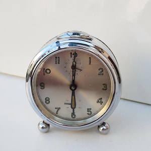 Può includere: Un orologio da tavolo argentato con quadrante bianco e lancette nere. L'orologio è rotondo e impostato alle 7:00. Ha una piccola lancetta dei secondi e un piccolo quadrante per impostare la sveglia.