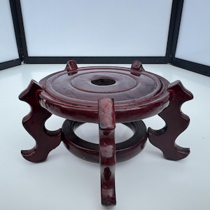May include: A dark red, wooden decorative stand with a circular top and three curved legs. The stand has a hole in the centre of the top and a ring-shaped base. The stand is likely used to display a decorative object.