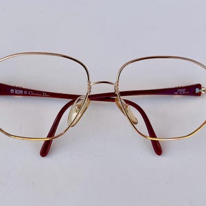 Op de afbeelding: Goudkleurige bril met een afgerond rechthoekig montuur. De veren en neuspads zijn diep bordeauxrood. De merknaam "Christian Dior" is zichtbaar op de poot.