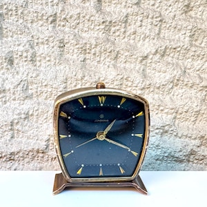 May include: A vintage Junghans table clock with a trapezoidal gold-tone metal frame and a black face. The clock features gold-colored triangular hour markers, thin gold hands, and the brand name Junghans printed in white on the center of the dial.