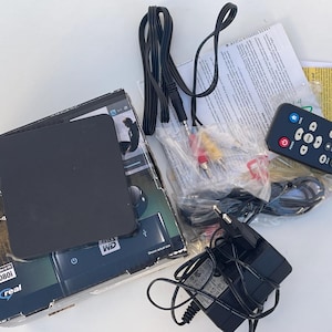 Op de afbeelding: Een zwarte WD TV Media Player-doos met een zwarte mediaspeler, afstandsbediening, kabels en voedingsadapter. De doos heeft het WD-logo en de tekst "WD TV Media Player" en "Play DVD-q".
