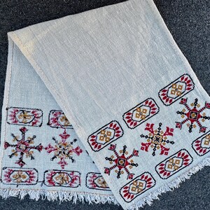 Può includere: Tessuto grigio chiaro con intricati motivi ricamati in rosso, oro e nero. Il design presenta forme geometriche e motivi a forma di fiocco di neve lungo i bordi, con un bordo sfrangiato. Il tessuto è piegato.