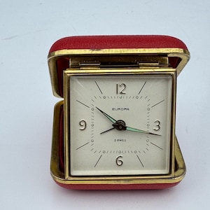 Op de afbeelding: Een vintage Europa reisklok in een rode behuizing. De vierkante wijzerplaat met een goudkleurige rand heeft zwarte cijfers en wijzers, en de tekst "EUROPA" en "2 JEWELS". De klok is open en toont de tijd.