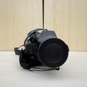 Könnte beinhalten: Eine schwarze Videokamera mit Objektivdeckel, Schultergurt und Handschlaufe. Die Kamera befindet sich auf einer hellen Oberfläche mit hellem Holzgrund.