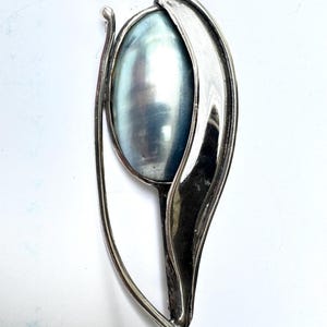 May include: A silver-toned brooch featuring a smooth, oval-shaped iridescent blue-grey stone set within a curved, polished metal frame that wraps around the side of the stone, creating an elongated, abstract shape with a pointed bottom end.