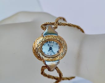 May include: A vintage Trifari wristwatch with a gold-tone oval face and a textured bezel. The watch has a white dial with black numerals and hands. The watch is attached to a gold-tone chain bracelet.