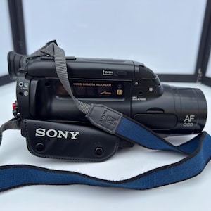 Peut inclure: Caméscope Sony noir avec une sangle bleue et grise. Le caméscope porte l'inscription "SONY" sur le côté et "VIDEO CAMERA RECORDER" sur le dessus. Le caméscope porte également l'inscription "AF CCD".